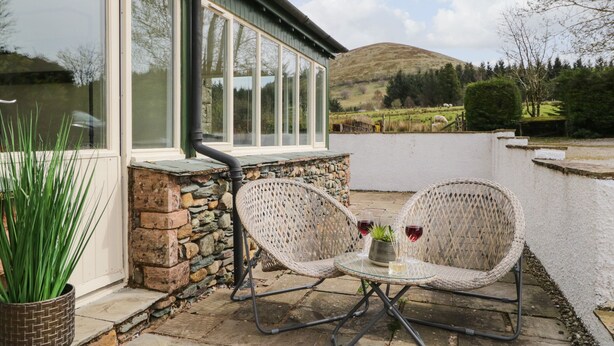 A patio with wicker chairs and a table, a stone wall, and a view of a hill and trees. - Glen Cottage (Penrith)