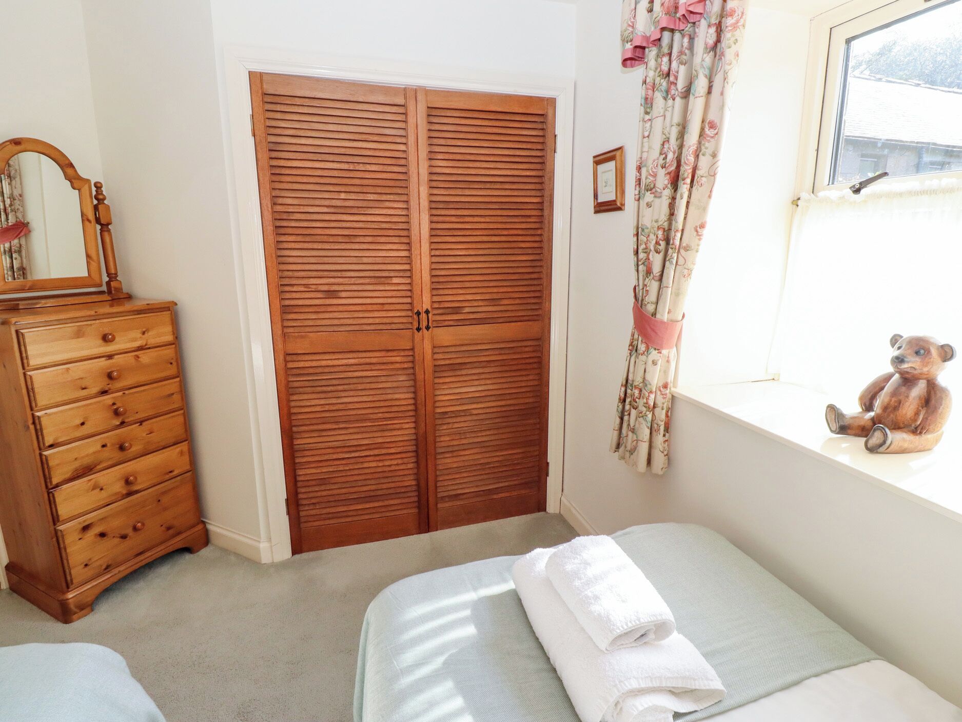 A bedroom with a wooden wardrobe, a dresser, a bed with a white towel, a window with floral curtains, and a teddy bear on the windowsill.