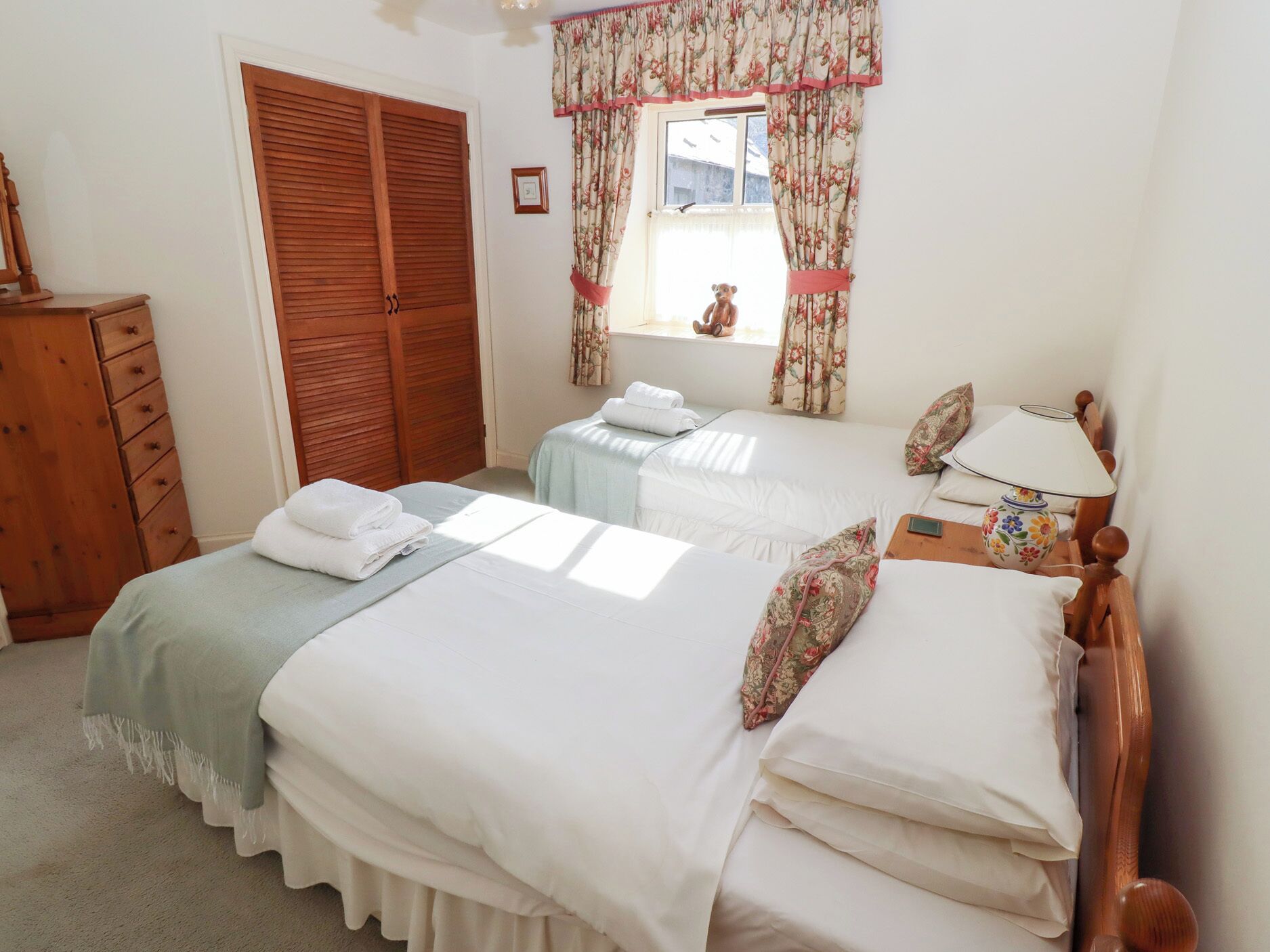 A bedroom with two beds, a wooden wardrobe, a window with floral curtains, and a bedside table with a lamp.