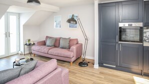 Ein modernes Wohnzimmer mit einem rosa Sofa, einem grauen Sofa, einer schwarzen Stehlampe und einer Küche mit dunklen Schränken und einem eingebauten Backofen.