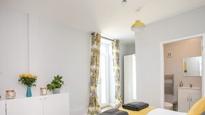 Ein Schlafzimmer mit Bett, Nachttisch mit Blumenvase, Spiegel und einem durchgehenden Türrahmen sichtbaren Badezimmer.