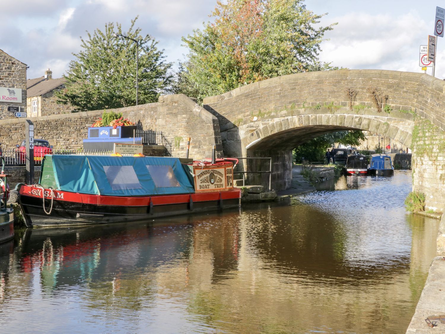 A canal with a stone bridge, a covered boat docked at the side, and a sign for a boat trip service.