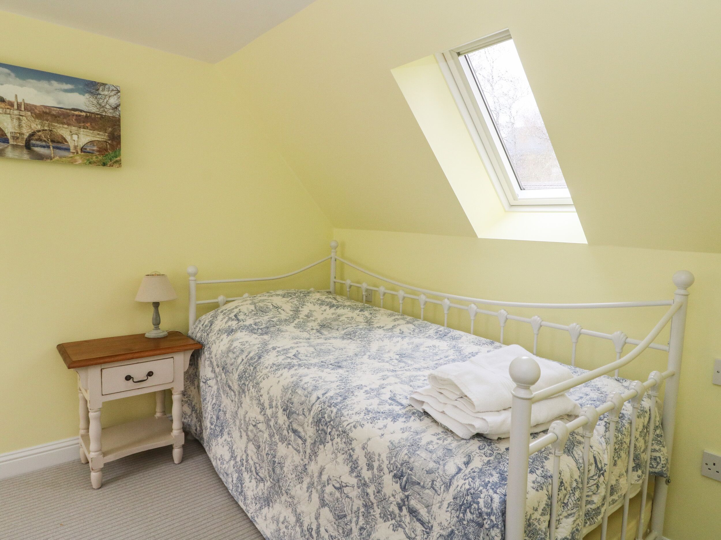 A bedroom with a single bed, a nightstand, a lamp, a painting on the wall, and a skylight.