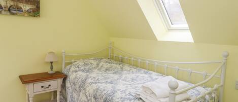 A bedroom with a single bed, a nightstand, a lamp, a painting on the wall, and a skylight.