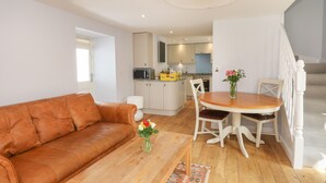 A modern living room with a brown leather sofa, a wooden coffee table, and a dining table with white chairs. A kitchen is visible in the background with white cabinets and a microwave. - Puccini (Pitlochry)