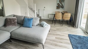 A modern living room with a grey sectional sofa, a staircase, a dining area with wicker chairs, and a large abstract painting on the wall. - 3 Parc Delfryn (Brynteg)