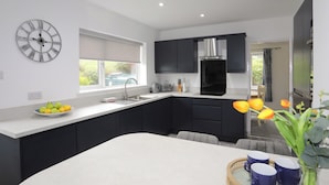A modern kitchen with dark cabinets, a white countertop, a built-in stove, and a large window. - Bwthyn Y Bae (Holyhead)