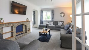 A living room with a stone fireplace, grey sofas, a coffee table, and a TV mounted on the wall. - Bwthyn Y Bae (Holyhead)