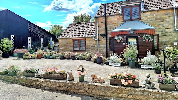 A stone building with a tiled roof, a sign reading "Farmyard Cottage Parking," and various potted plants and garden decorations. - Farm Yard Cottage (Pickering)