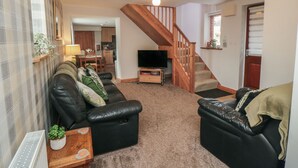 A living room with a black leather sofa, a TV, and a wooden staircase. - Farm Yard Cottage (Pickering)