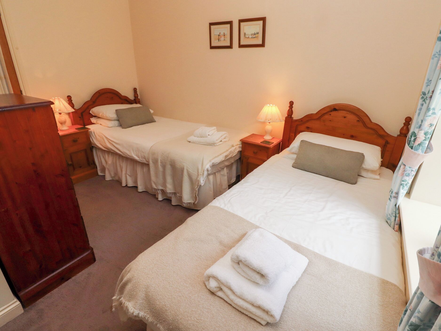 A hotel room with two single beds, each with a wooden headboard, a bedside table with a lamp, and a small drawer. The room has a carpeted floor and plain walls.