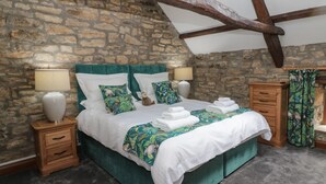Un dormitorio con pared de piedra, viga de techo de madera, cama con ropa de cama verde, dos mesitas de noche y un tocador.