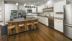 A modern kitchen with white cabinetry, stainless steel appliances, a wooden island with bar stools, and a black trash can. - Locust Estates Luxury Home – Downtown Windsor (Windsor)