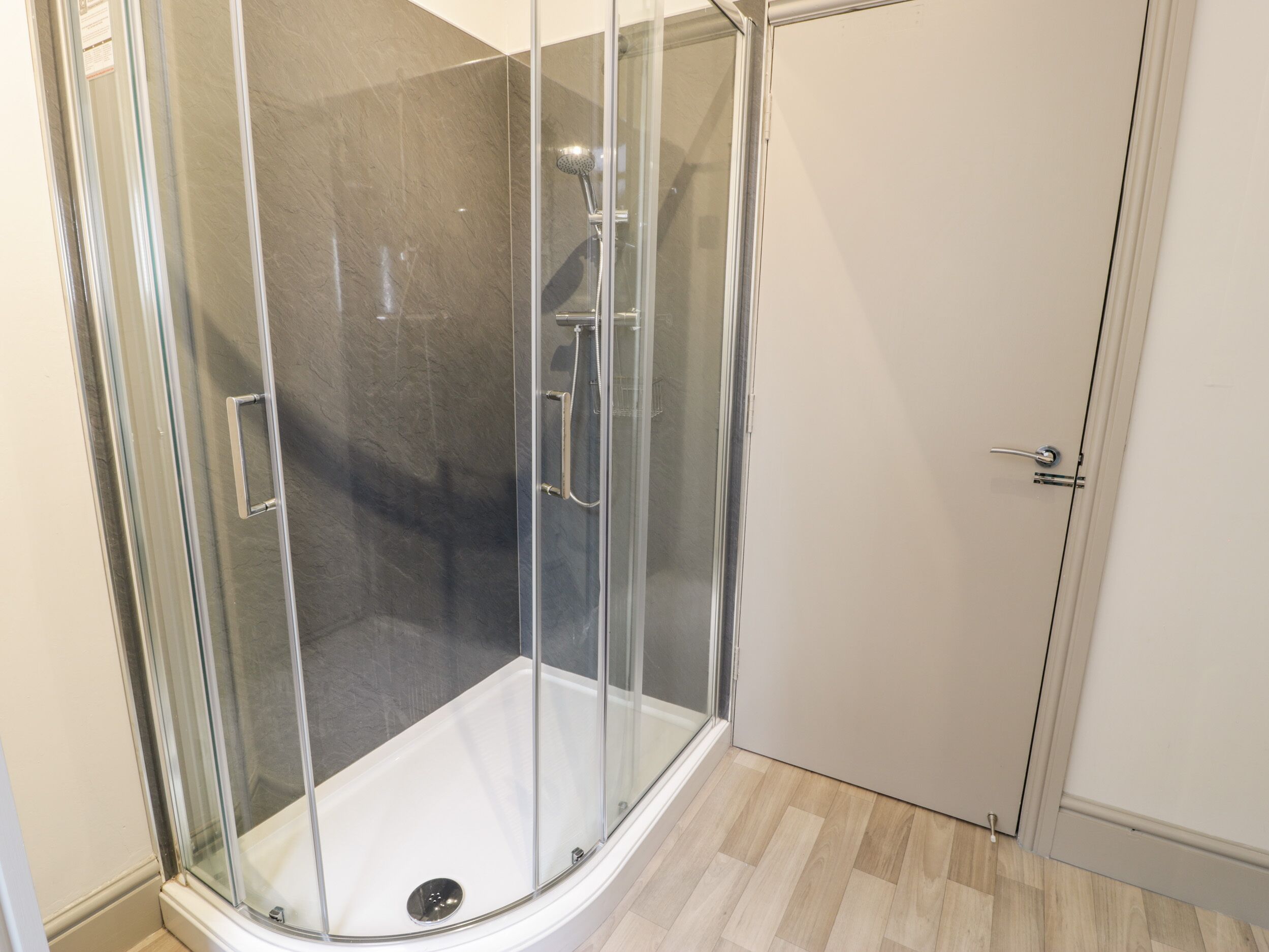Cabina de ducha con paredes de vidrio, suelo blanco y puerta a la derecha.
