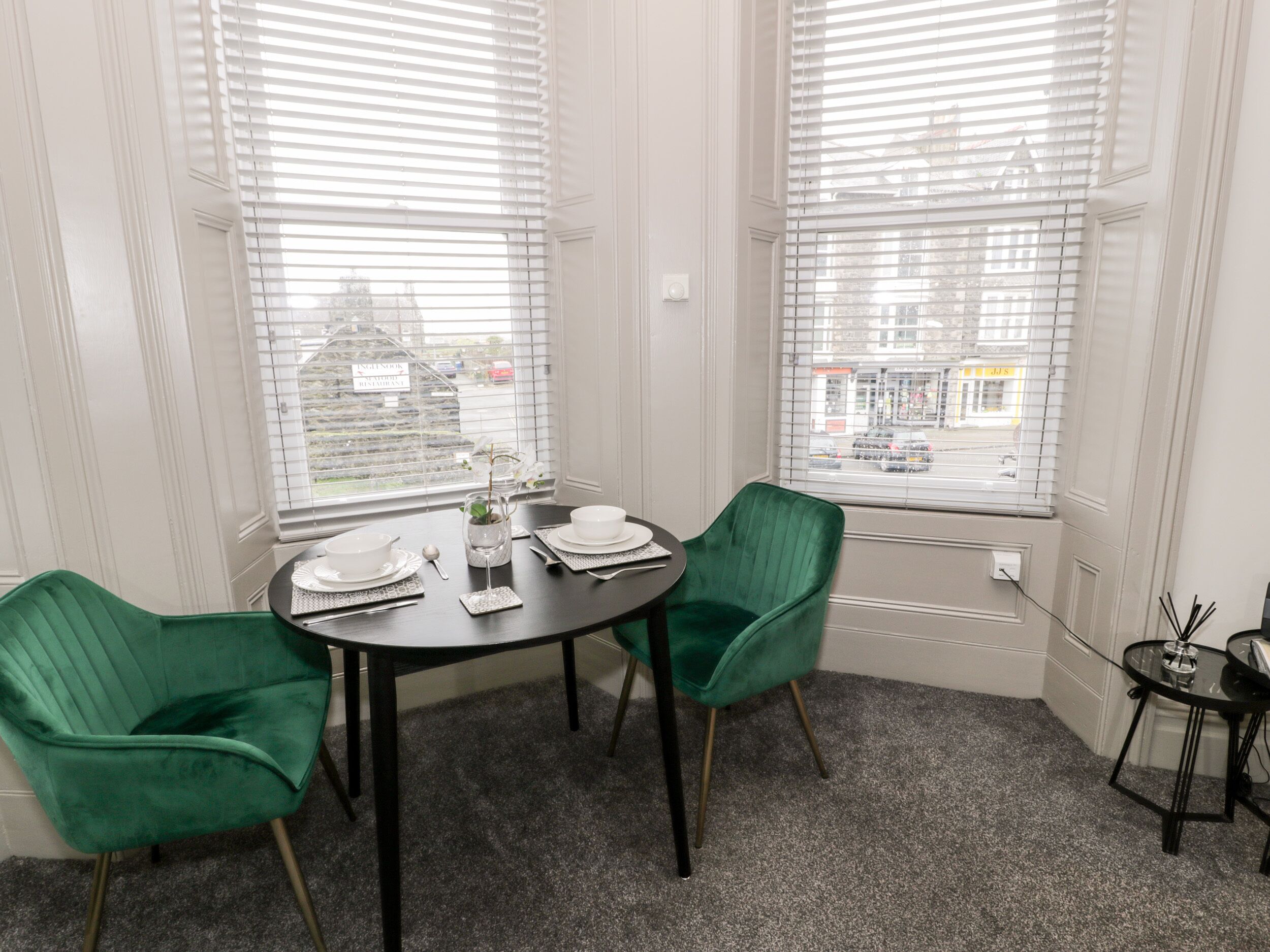 A dining area with a round table, two green chairs, and a small side table with a vase and a candle.