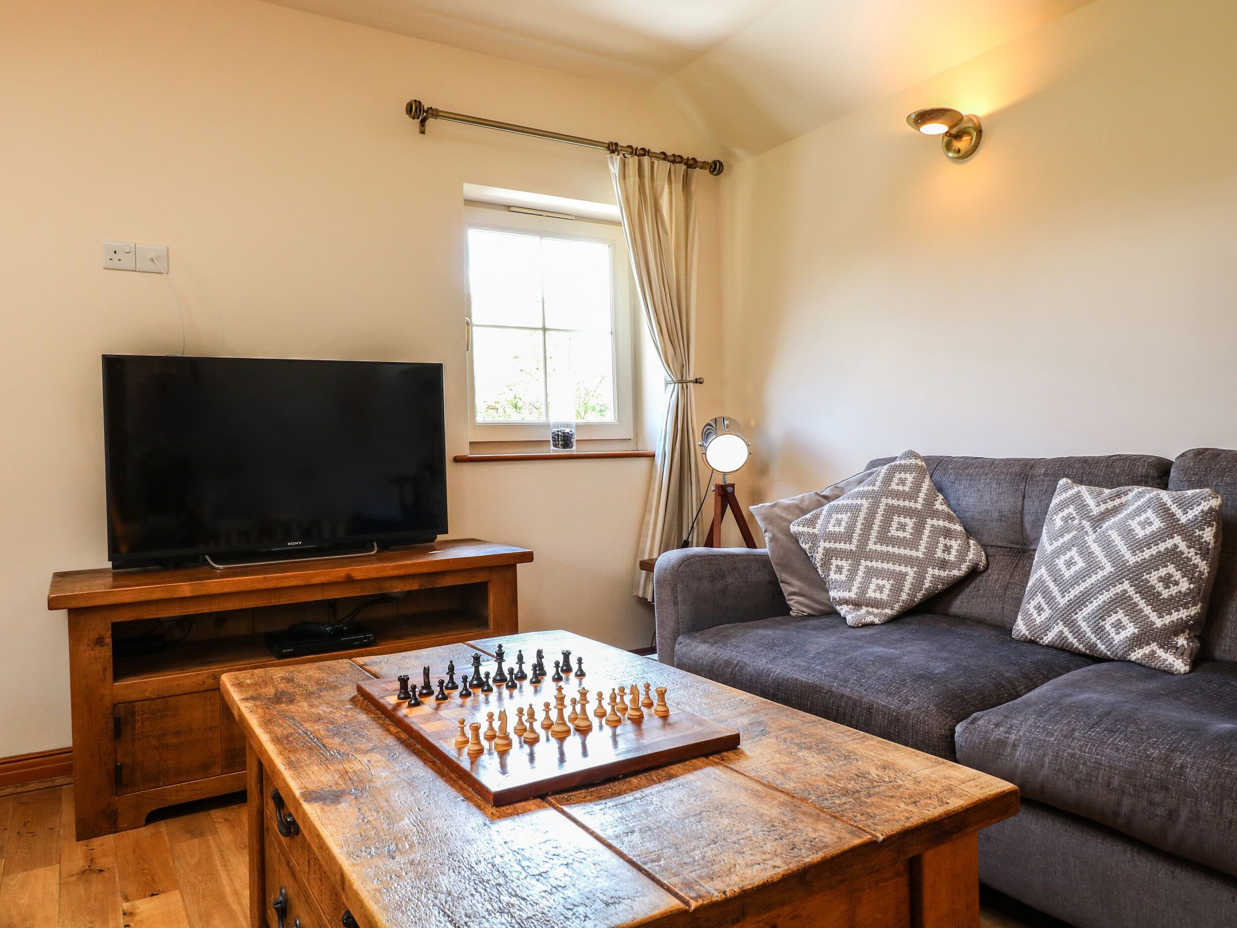 A living room with a flat-screen TV, a wooden coffee table with a chessboard, a grey sofa with patterned cushions, and a window with curtains.