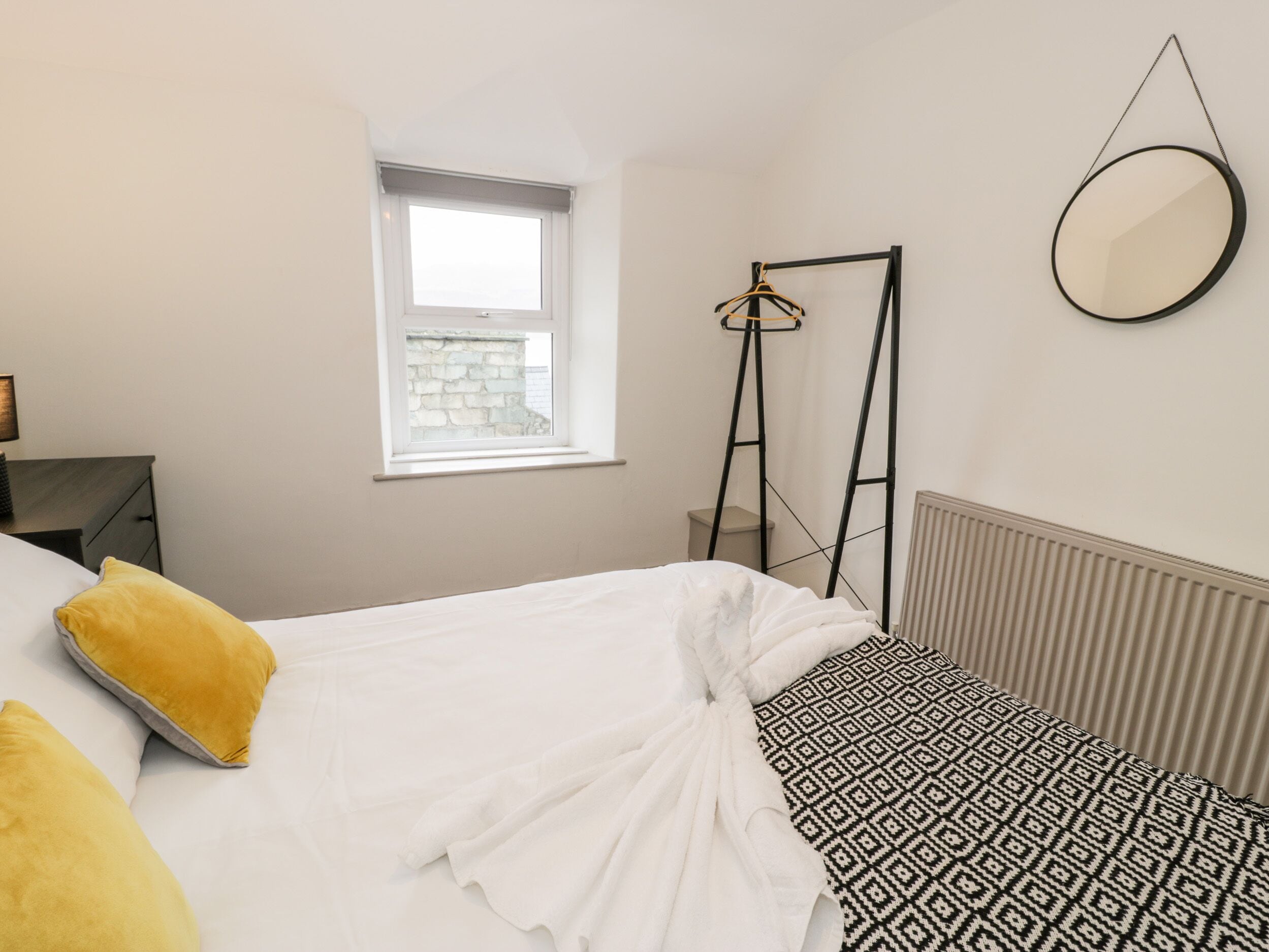 Ein Schlafzimmer mit Bett, Fenster, Heizkörper und einem hängenden Spiegel.