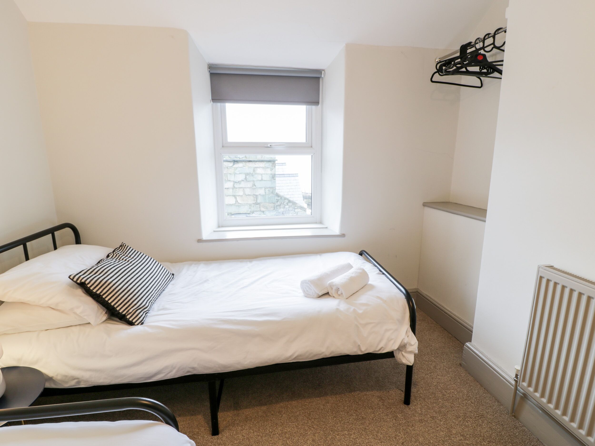 A single bed with white linens, a striped pillow, and a rolled towel. A window with a view of a stone building is above the bed.