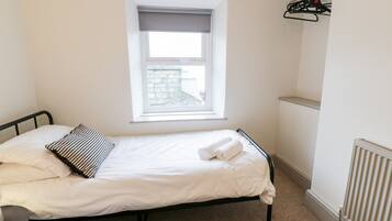 A single bed with white linens, a striped pillow, and a rolled towel. A window with a view of a stone building is above the bed.