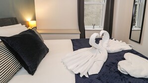 Una habitació d'hotel amb un llit ben fet, una làmpada de nit, una finestra amb cortines i una estanteta penjada a la paret amb una planta.