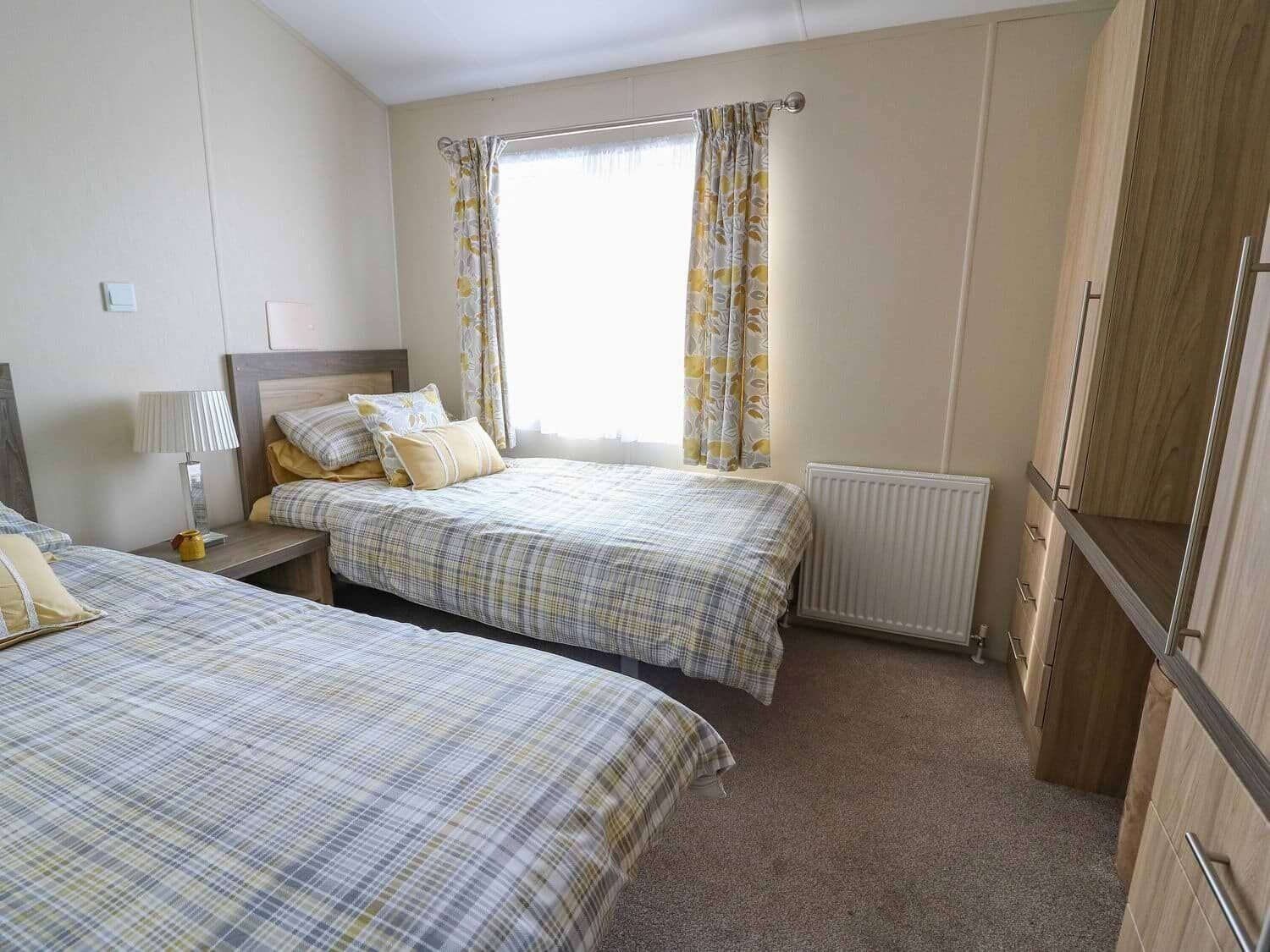 A bedroom with two beds, a nightstand, a lamp, a window with curtains, a radiator, and wooden wardrobe.