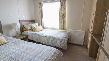 A bedroom with two beds, a nightstand, a lamp, a window with curtains, a radiator, and wooden wardrobe.