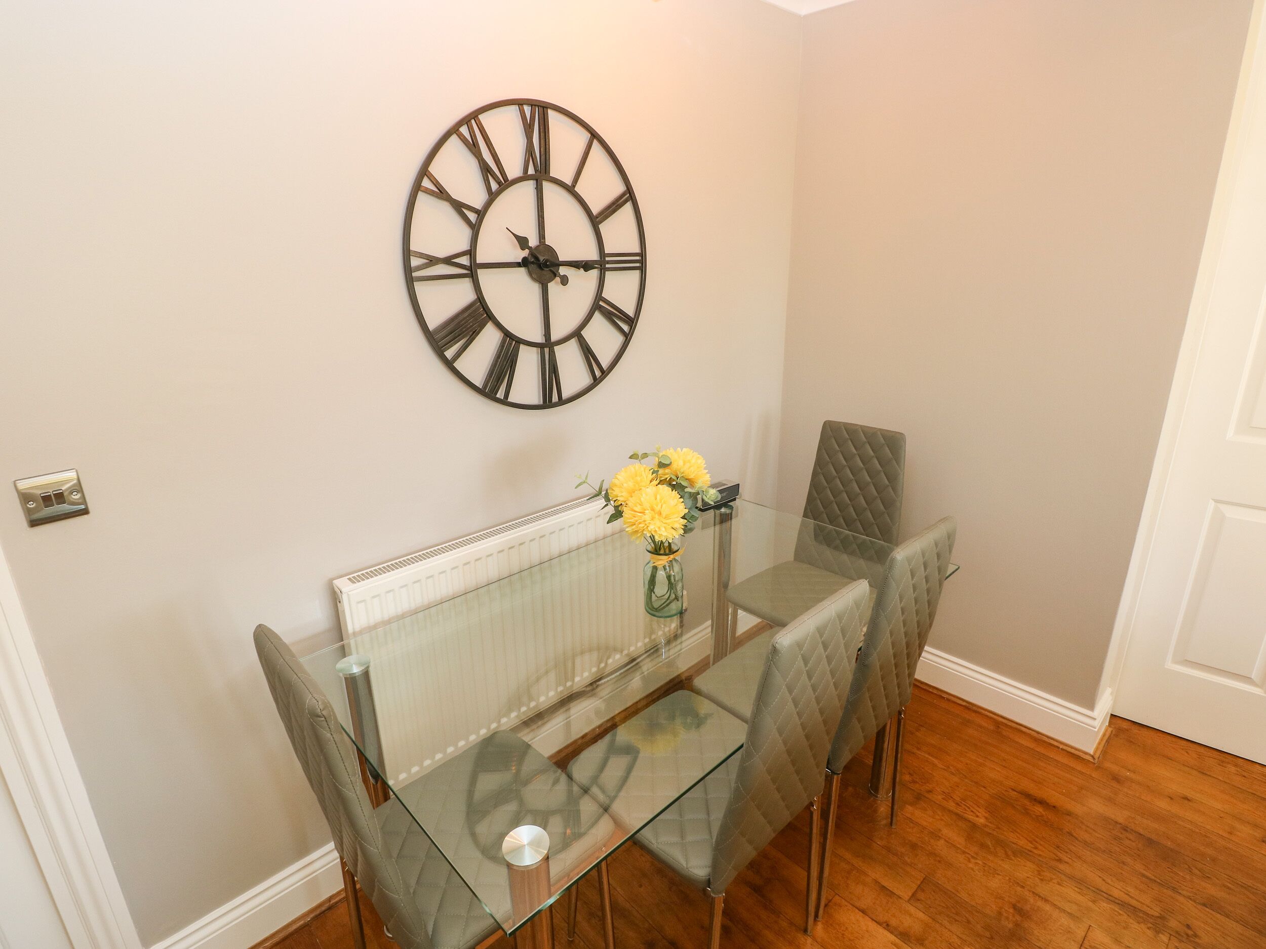 Un comedor con mesa de vidrio, dos sillas, un florero con flores y un reloj de pared.