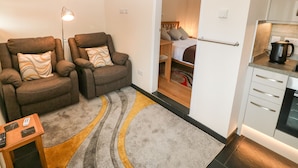 Un espacio habitable pequeño con dos sillones reclinables, una mesita, una zona de cocina con hervidor y una habitación visible a través de una puerta abierta.