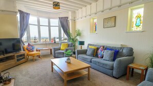 Una sala de estar con sofá, sillones, una mesa de centro, televisor y vistas a la playa a través de amplios ventanales.