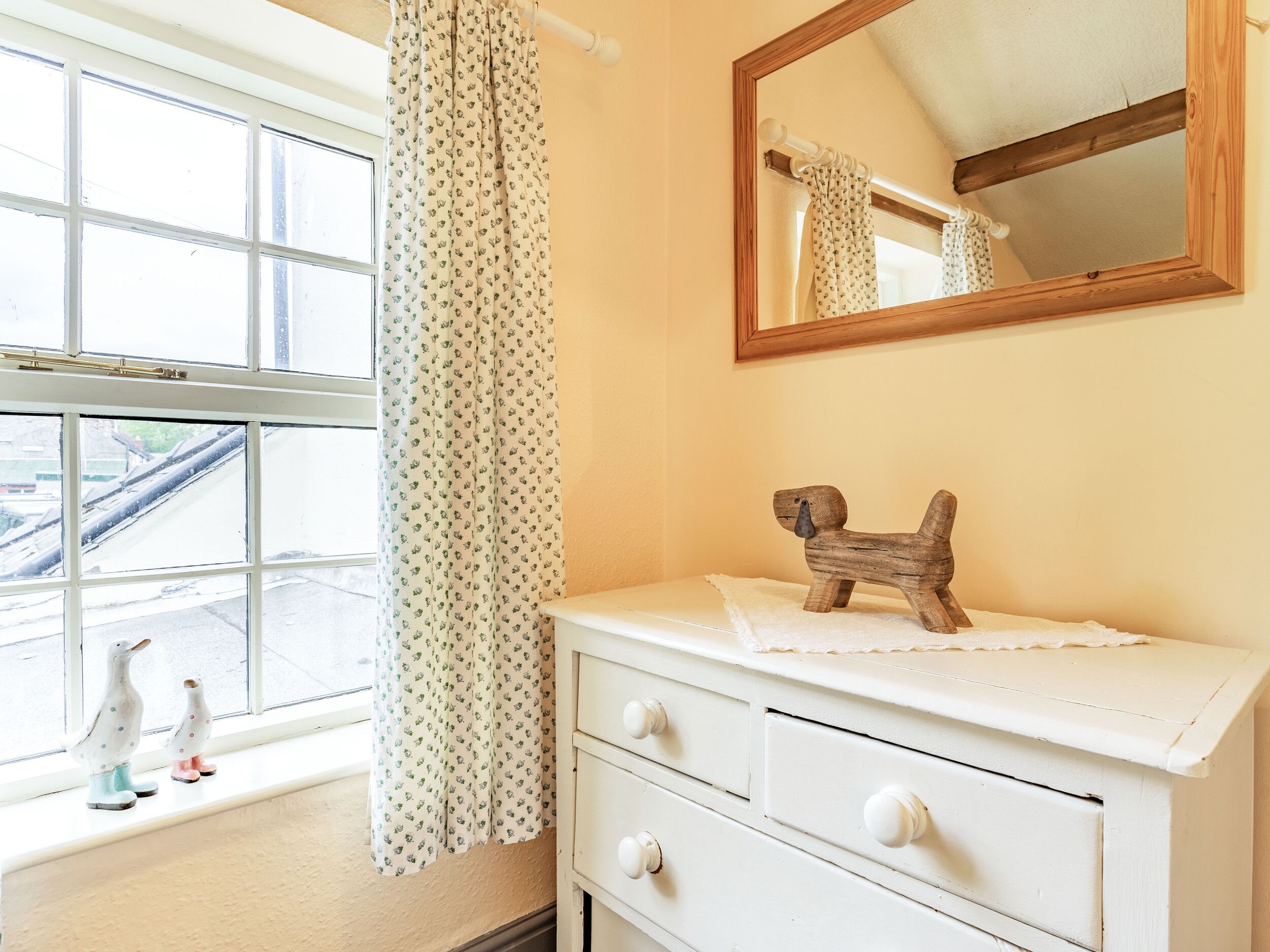 A bathroom with a white dresser, a wooden dog figurine, a mirror, and a window with a view of the street.