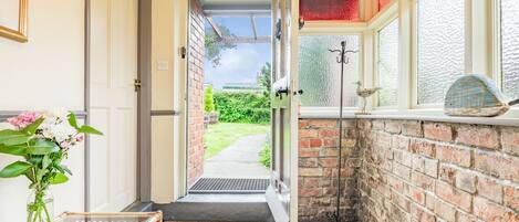 A narrow hallway with a door leading to a garden, a small table with a vase of flowers, and a window with a decorative bird.