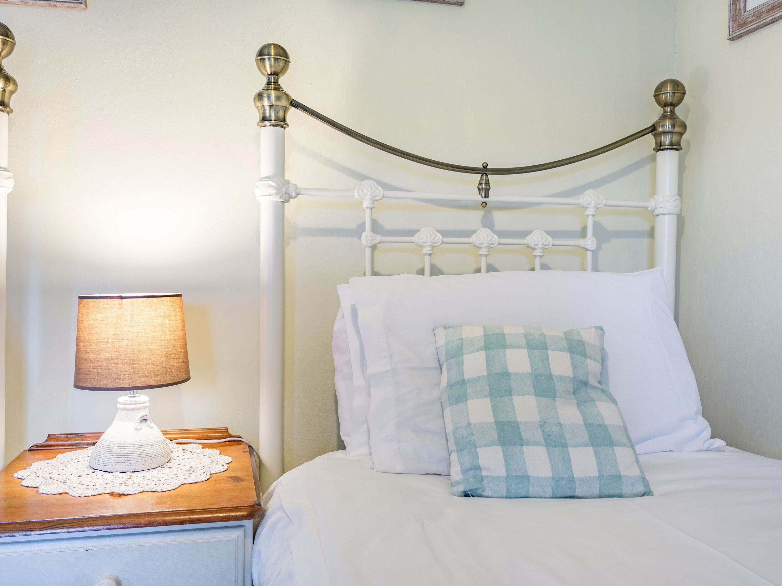 A neatly made bed with white bedding and a checkered pillow, a bedside table with a lamp, and a white metal headboard.