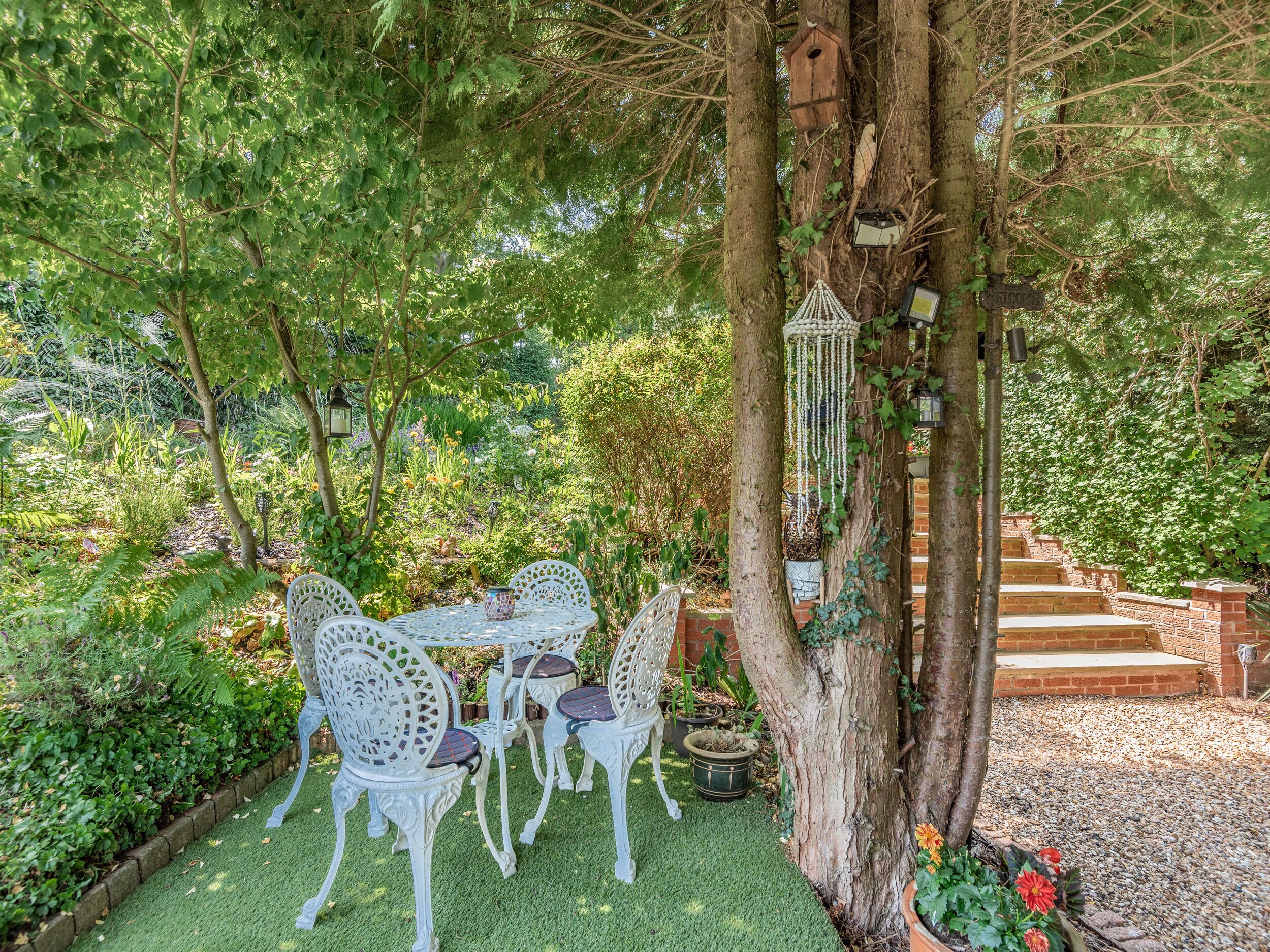 An outdoor seating area with white chairs and a table, surrounded by lush greenery and a tree with a birdhouse.