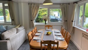 Een eetgedeelte met een houten tafel voor vier personen, omringd door houten stoelen. Een bank staat links, en een raam met gordijnen is zichtbaar.