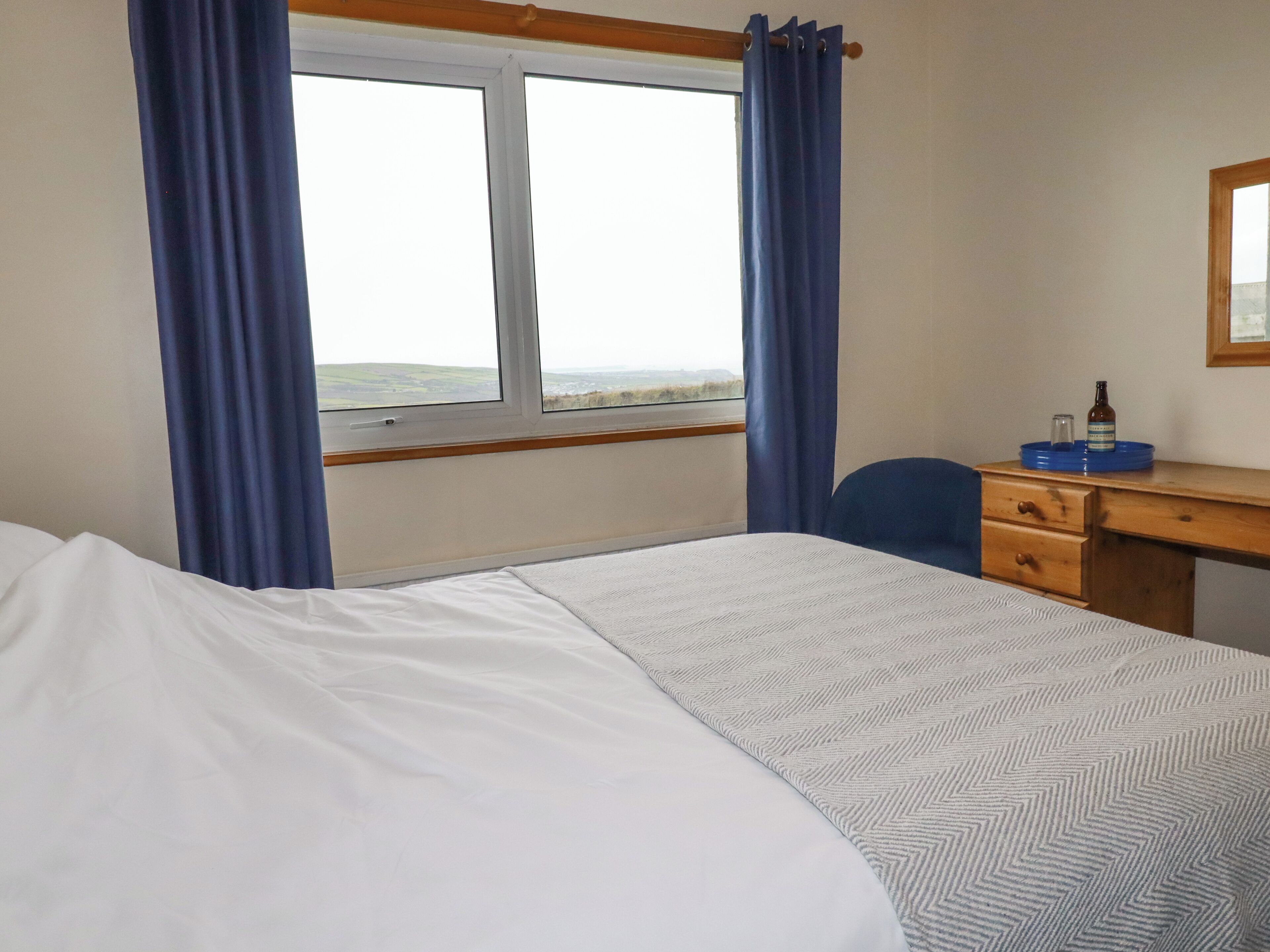 Quarto de hotel com cama, escrivaninha, cadeira, janela com cortinas e vista para o campo.