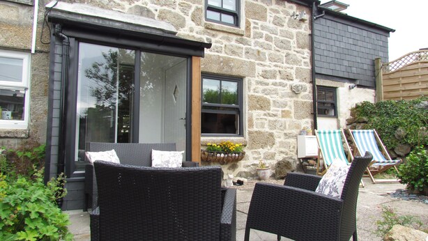 A stone building with a large glass door, wicker furniture, and a small garden area with flowers and a wooden fence. - Trenwith Bridge Cottage (St Ives)