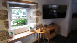 A room with a stone wall, a window with plants, a wooden bench, a small table with a bowl, and a TV mounted on the wall. - Trenwith Bridge Cottage (St Ives)