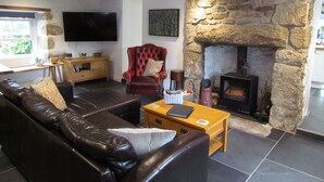 A living room with a stone fireplace, leather sofa, and a flat-screen TV mounted on the wall. - Trenwith Bridge Cottage (St Ives)