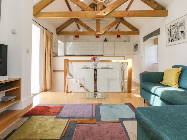 Ein modernes Wohnzimmer mit Holzboden, einem grünen Sofa, einem farbenfrohen Teppich und einer Küchenzeile im Hintergrund.