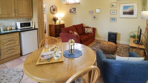 A cozy living room with a red sofa, a wooden dining table with a tray of food, and a kitchen area with wooden cabinets and a microwave. - The Garden Lodge (Bridgnorth)