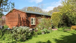 A wooden cabin with a small window, surrounded by a well-maintained garden and trees. - The Garden Lodge (Bridgnorth)