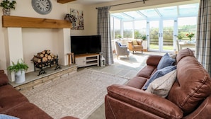 A spacious living room with a large brown leather sofa, a television on a wooden stand, a fireplace, and a large window offering a view of the outdoors. - West Reins (Barnard Castle)