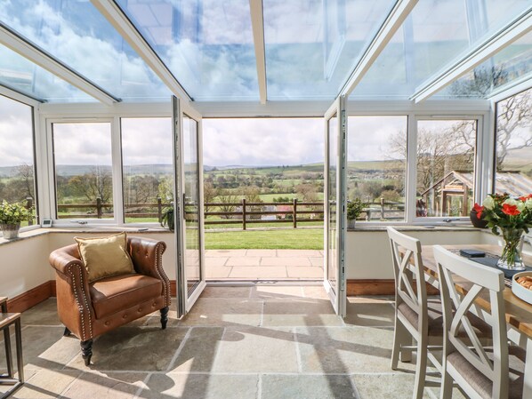 A conservatory with a view of a rural landscape, a leather sofa, and a dining table with chairs. - West Reins (Barnard Castle)