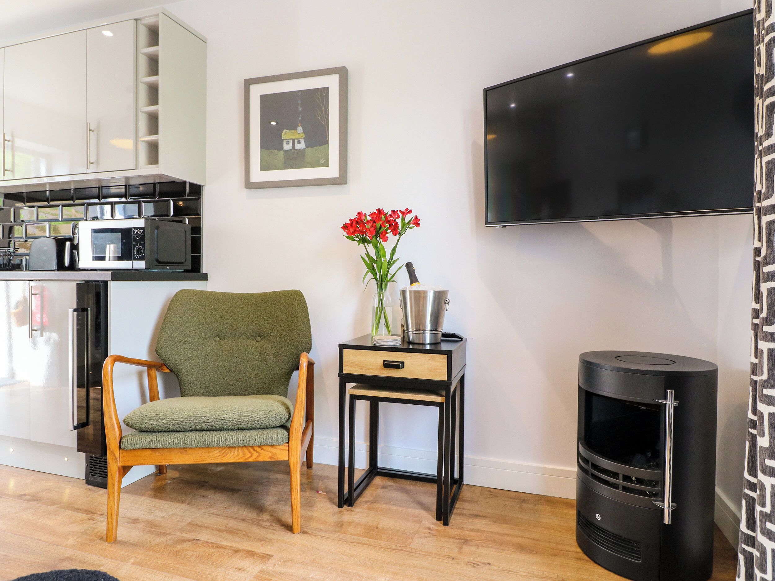 A modern living room with a green armchair, a small table with a vase of red flowers, a black fireplace, and a flat-screen TV mounted on the wall.