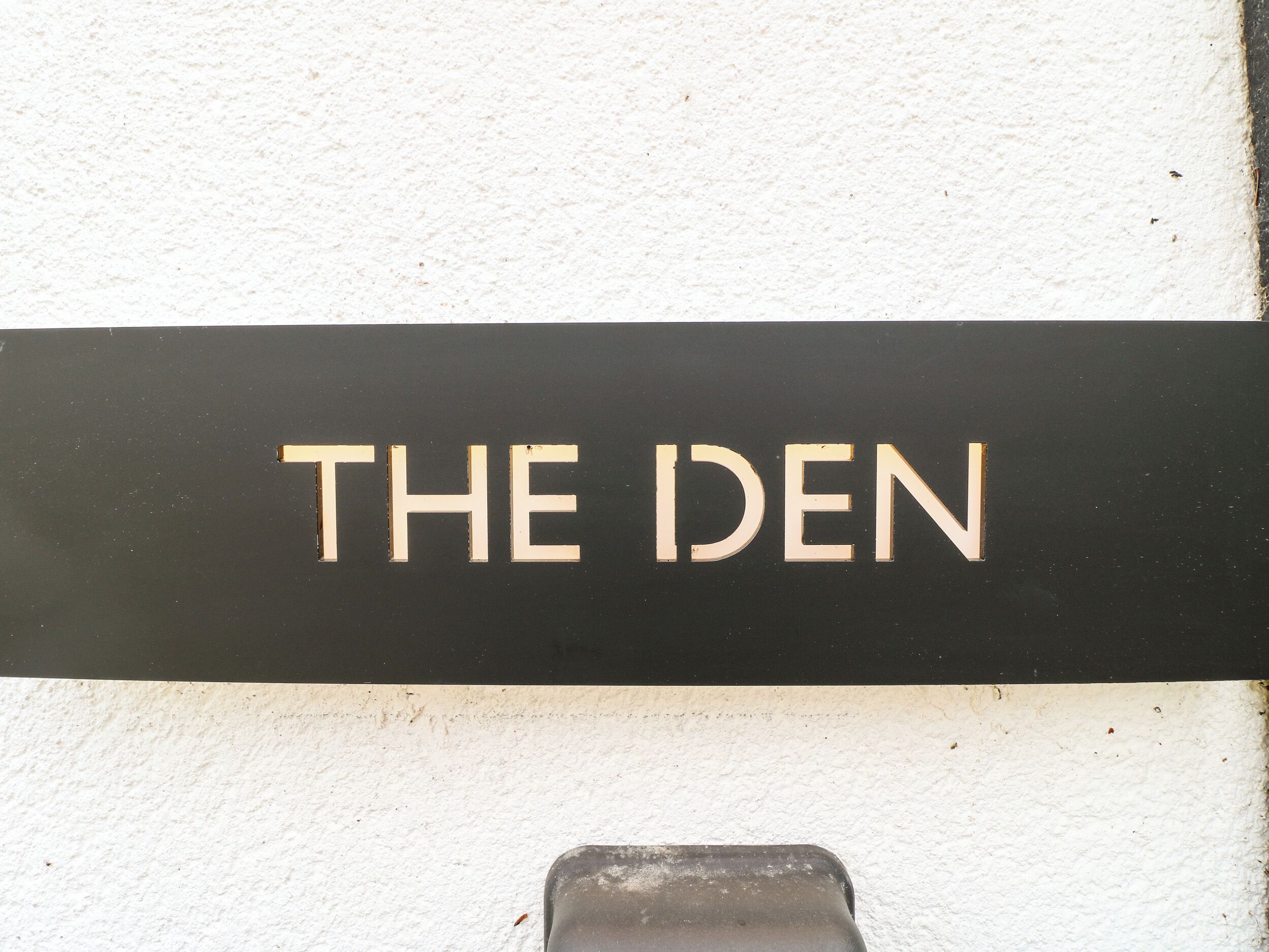 Un letrero negro con las palabras 'THE DEN' en letras blancas mayúsculas sobre una pared blanca.