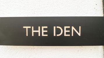 Un letrero negro con las palabras 'THE DEN' en letras blancas mayúsculas sobre una pared blanca.