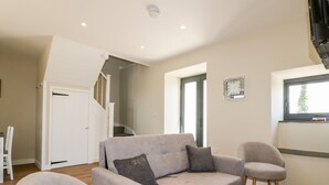 Un salon avec un canapé gris, deux fauteuils, un plancher en bois, un escalier et une fenêtre avec des stores.