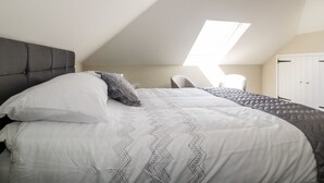 Une chambre avec un plafond en pente, un lit doté d’une tête de lit grise, une chaise grise et une armoire.