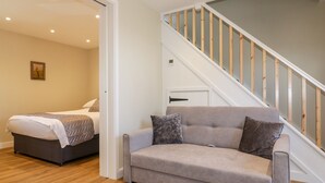 A modern living room with a grey sofa, a wooden staircase, and a bed in the adjacent room.