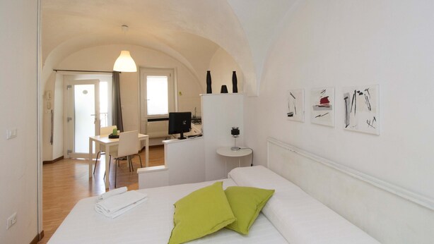 Ein Hotelzimmer mit Bett, Schreibtisch und Stuhl. Es gibt ein Fenster, eine Tür und eine Wand mit Kunstwerken.